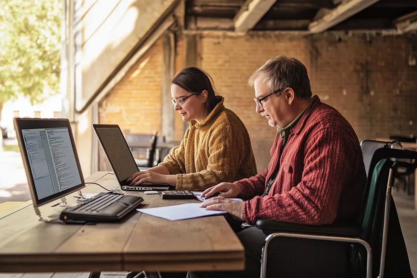 Bild 2 im Slider: Eine Frau und ein Mann sitzen an einem Tisch und arbeiten an zwei Computern. Der Mann sitzt in einem Rollstuhl und ist sehbehindert.